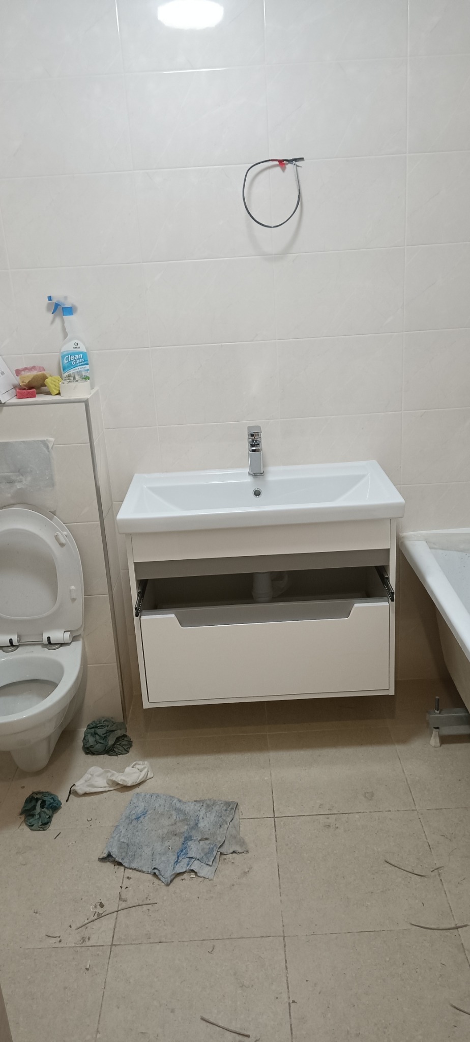 Biała umywalka z szafką w łazience z otwartą szufladą, widoczny kran. Na podłodze leżą ścierki, a na ścianie wisi kabel. W tle płytki i fragment wanny.