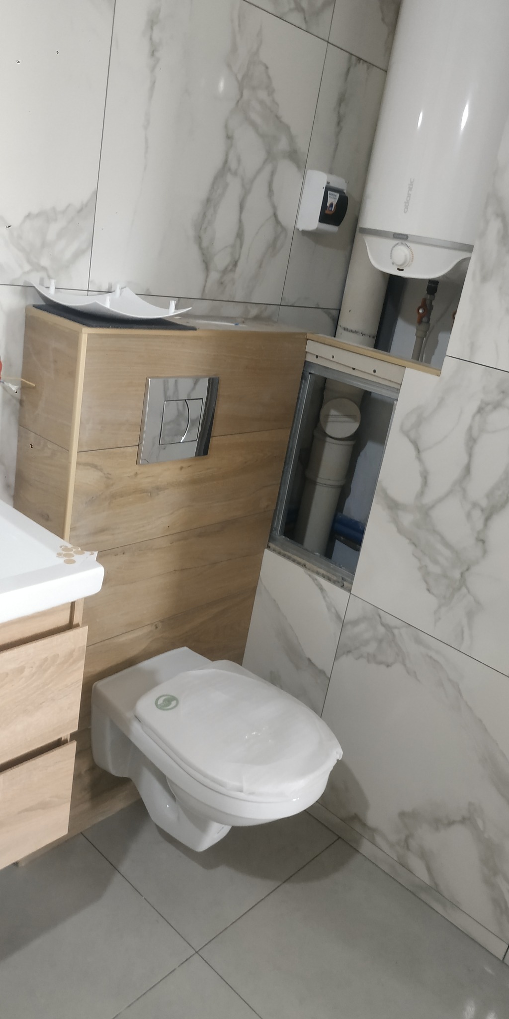 Biała toaleta podwieszana z drewnianą obudową i chromowanym przyciskiem spłukującym, na tle marmurowych płytek. Widoczny bojler i instalacja.