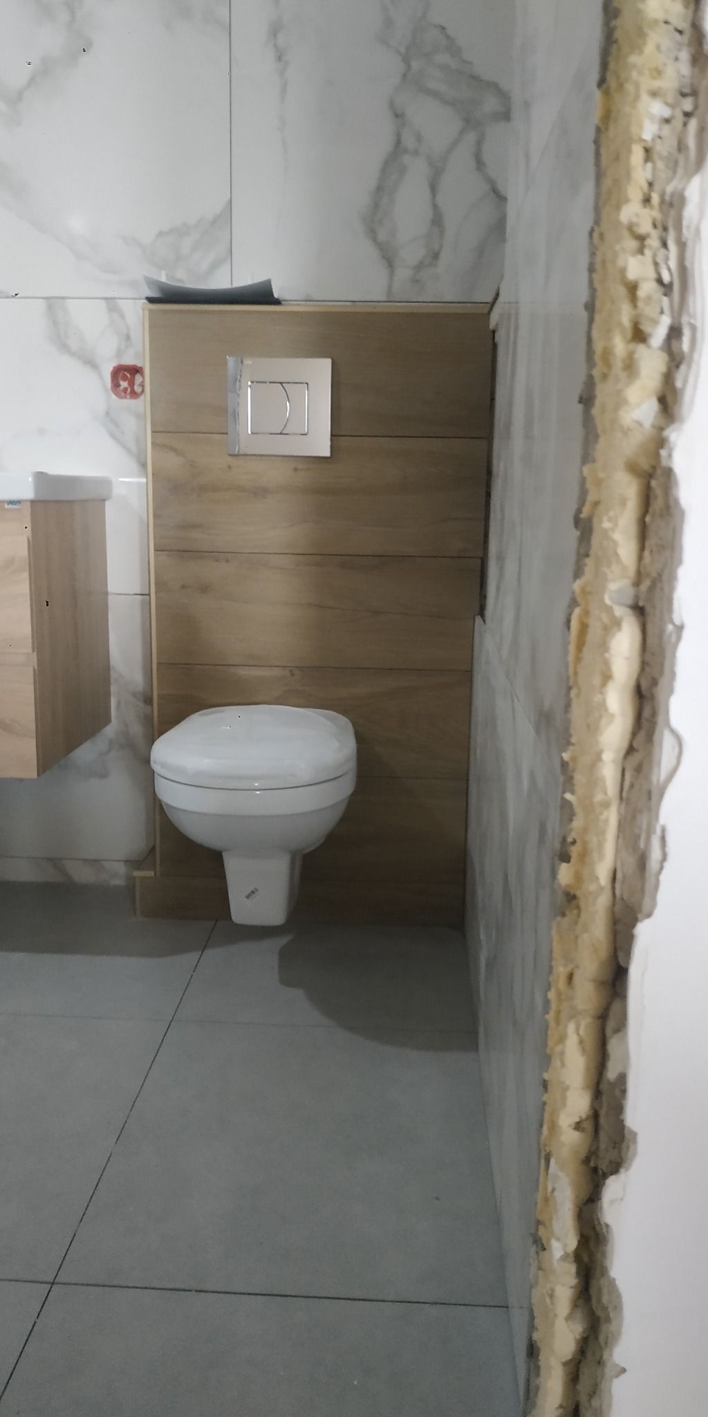 Biała toaleta wisząca ze srebrnym przyciskiem spłukującym na tle drewnopodobnej zabudowy, szare płytki podłogowe i marmurowe płytki ścienne. Widoczna krawędź ściany w trakcie remontu.