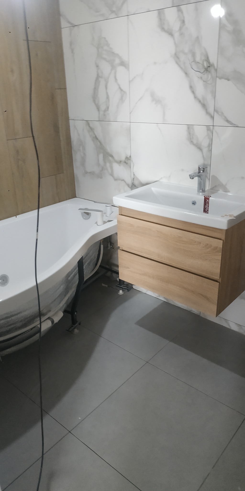 Nowoczesna łazienka w trakcie wykończenia: wanna z hydromasażem i umywalka z drewnianą szafką na tle marmurowych płytek. Szare płytki podłogowe.