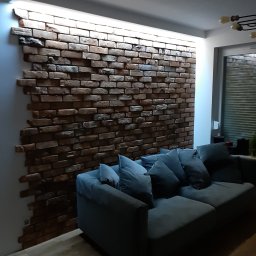 DOKŁADNI.PL SPÓŁKA Z OGRANICZONĄ ODPOWIEDZIALNOŚCIĄ - Ściana z cegły dekoracyjnej z podświetleniem LED w salonie, szara sofa z poduszkami. Widok na fragment okna i podłogę. Aranżacja wnętrza w stylu nowoczesnym.