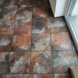 DOKŁADNI.PL SPÓŁKA Z OGRANICZONĄ ODPOWIEDZIALNOŚCIĄ - Rustykalne, kwadratowe płytki podłogowe w odcieniach brązu, szarości i bieli, ułożone przy oknie z widokiem na zieleń. Widoczna fuga i fragment progu.