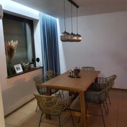 DOKŁADNI.PL SPÓŁKA Z OGRANICZONĄ ODPOWIEDZIALNOŚCIĄ - Jadalnia z drewnianym stołem i plecionymi krzesłami, oświetlona trzema wiszącymi lampami. Okno z zasłonami i dekoracjami. Ciepłe oświetlenie wnętrza.