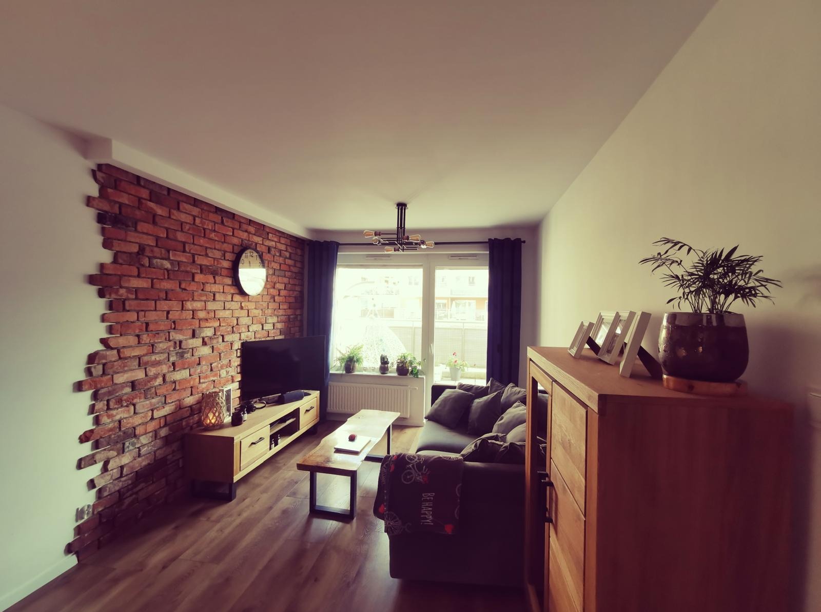 Salon z ceglaną ścianą, drewnianą podłogą i meblami w stylu skandynawskim. Widoczny telewizor, kanapa i roślina doniczkowa. Ściana z cegły częściowo pomalowana na biało.