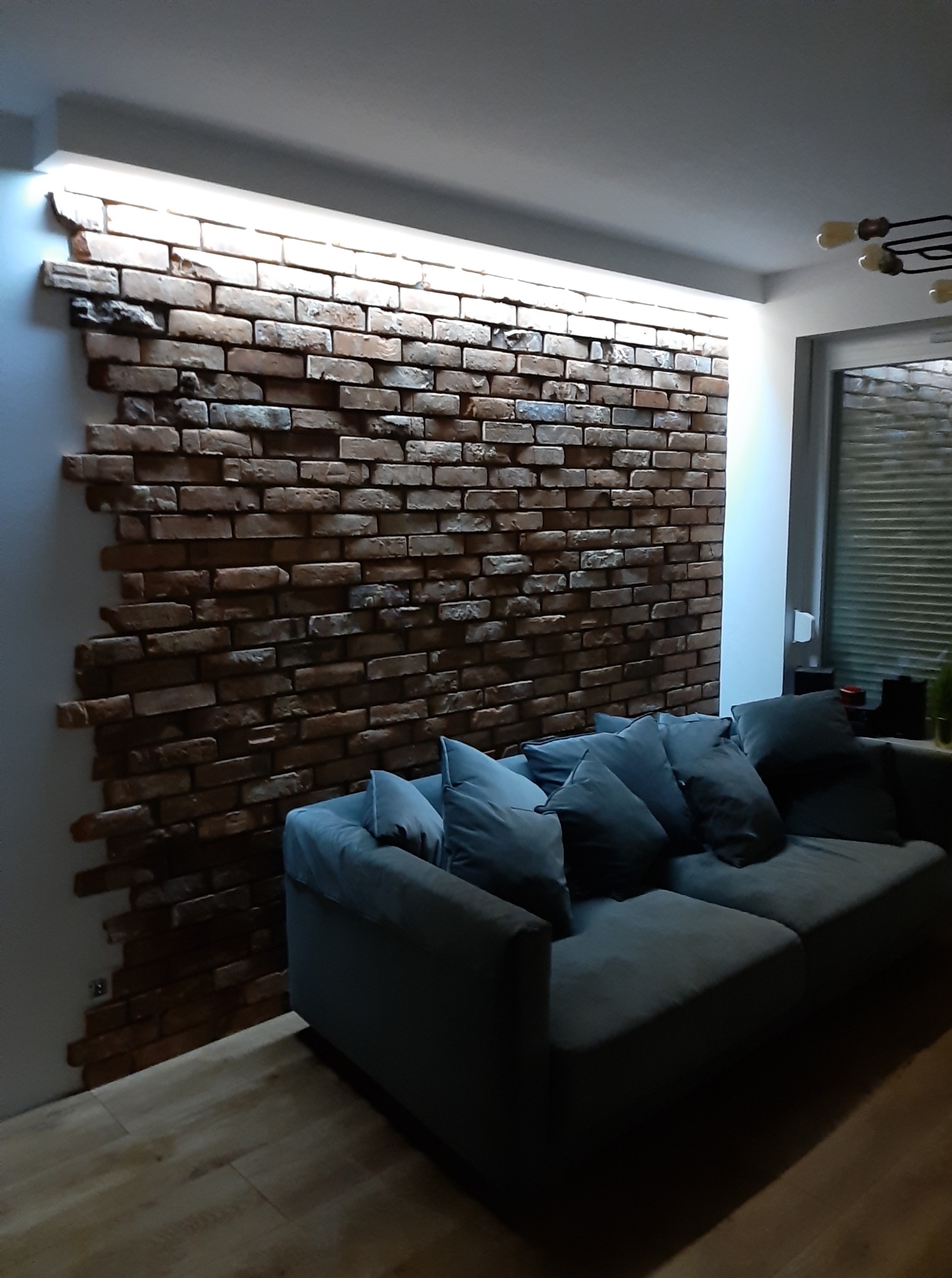 Ściana z cegły dekoracyjnej z podświetleniem LED w salonie, szara sofa z poduszkami. Widok na fragment okna i podłogę. Aranżacja wnętrza w stylu nowoczesnym.