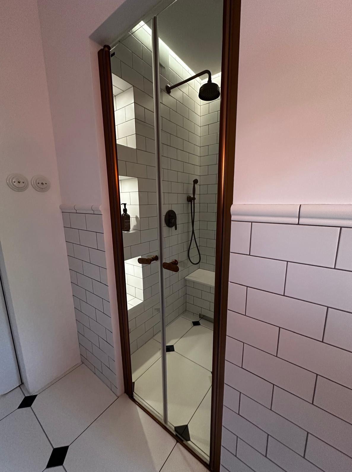 Nowoczesna kabina prysznicowa z brązowymi ramami i białymi płytkami w stylu retro, z półkami w ścianie i czarnymi akcentami na podłodze.