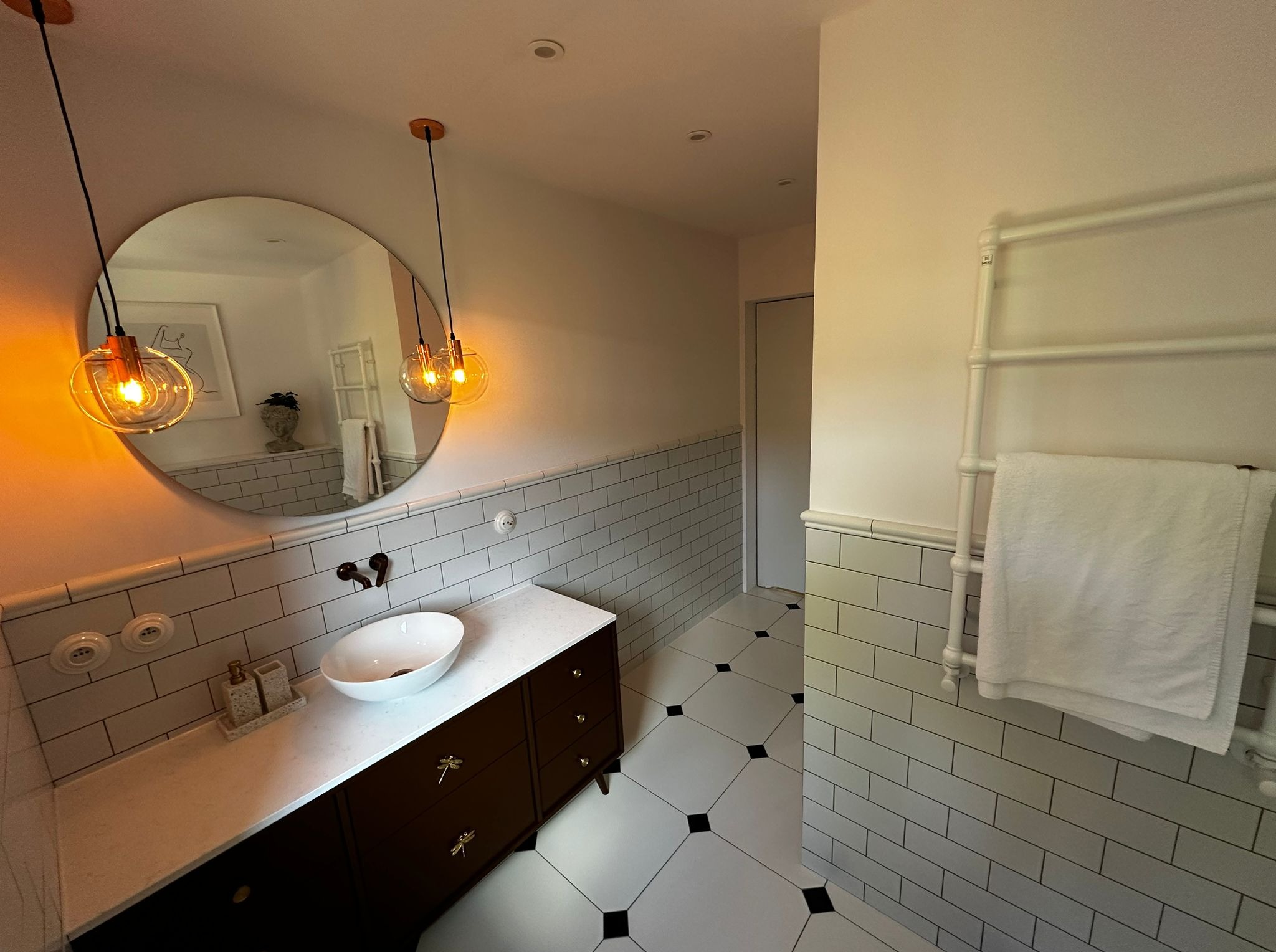 Biała łazienka z umywalką nablatową, brązową szafką z motywem ważki, okrągłym lustrem i lampami. Płytki na ścianach i podłodze w stylu retro.