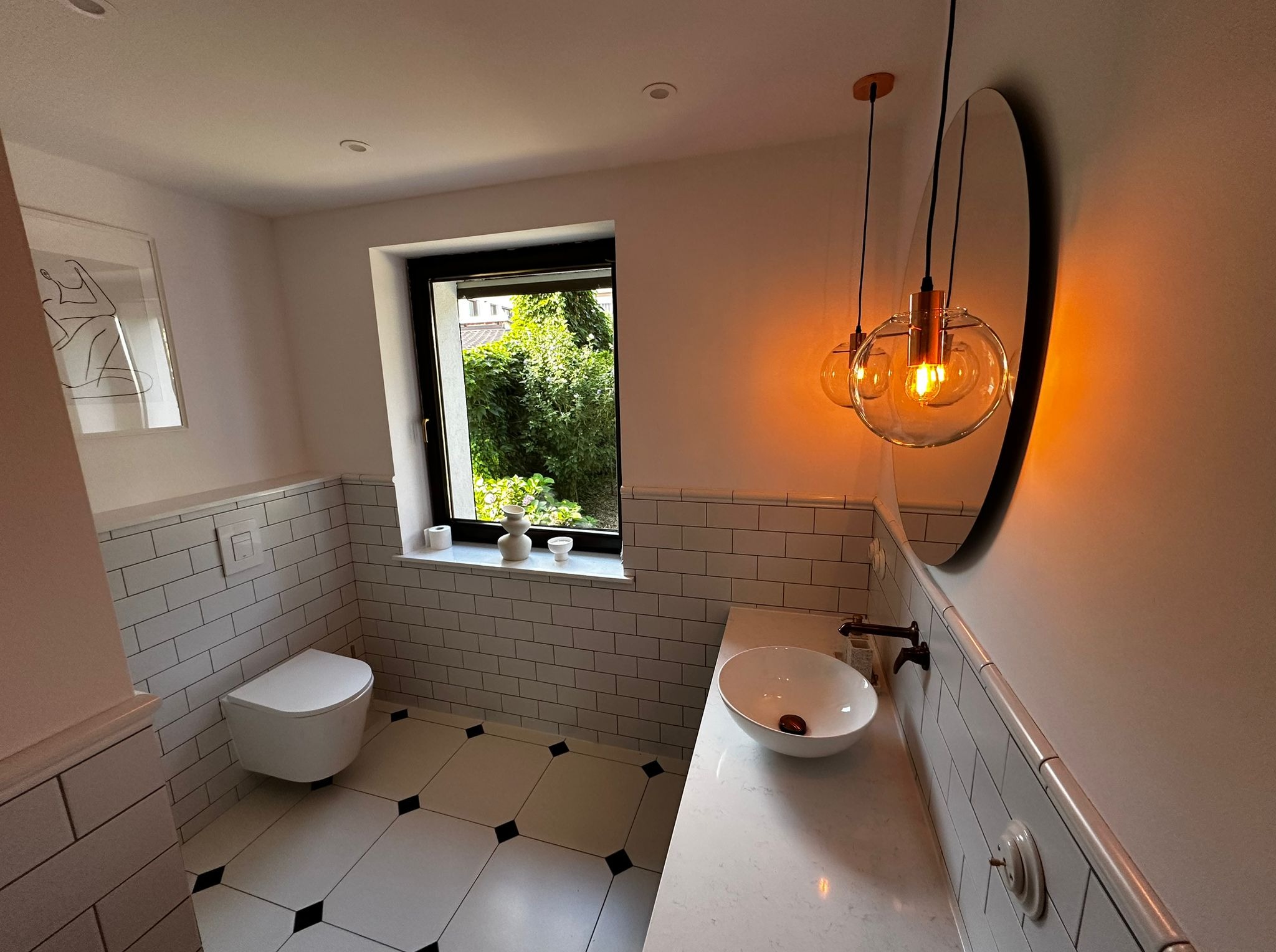 Nowoczesna łazienka z białą armaturą, czarnymi akcentami i oświetleniem w stylu loft. Widok na okno z zielenią.