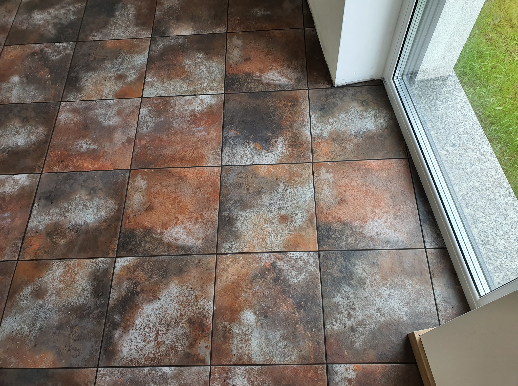 Rustykalne, kwadratowe płytki podłogowe w odcieniach brązu, szarości i bieli, ułożone przy oknie z widokiem na zieleń. Widoczna fuga i fragment progu.