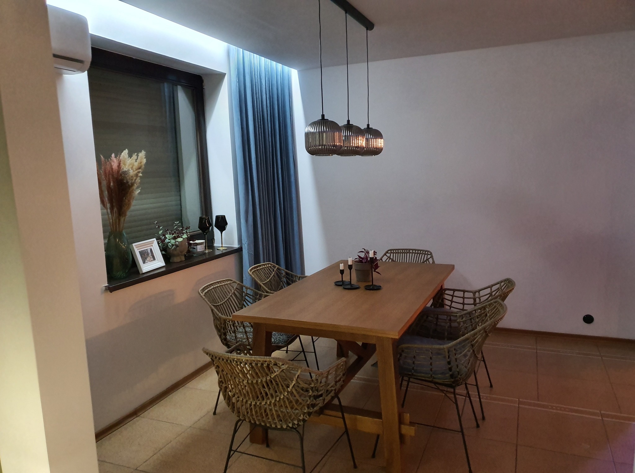 Jadalnia z drewnianym stołem i plecionymi krzesłami, oświetlona trzema wiszącymi lampami. Okno z zasłonami i dekoracjami. Ciepłe oświetlenie wnętrza.