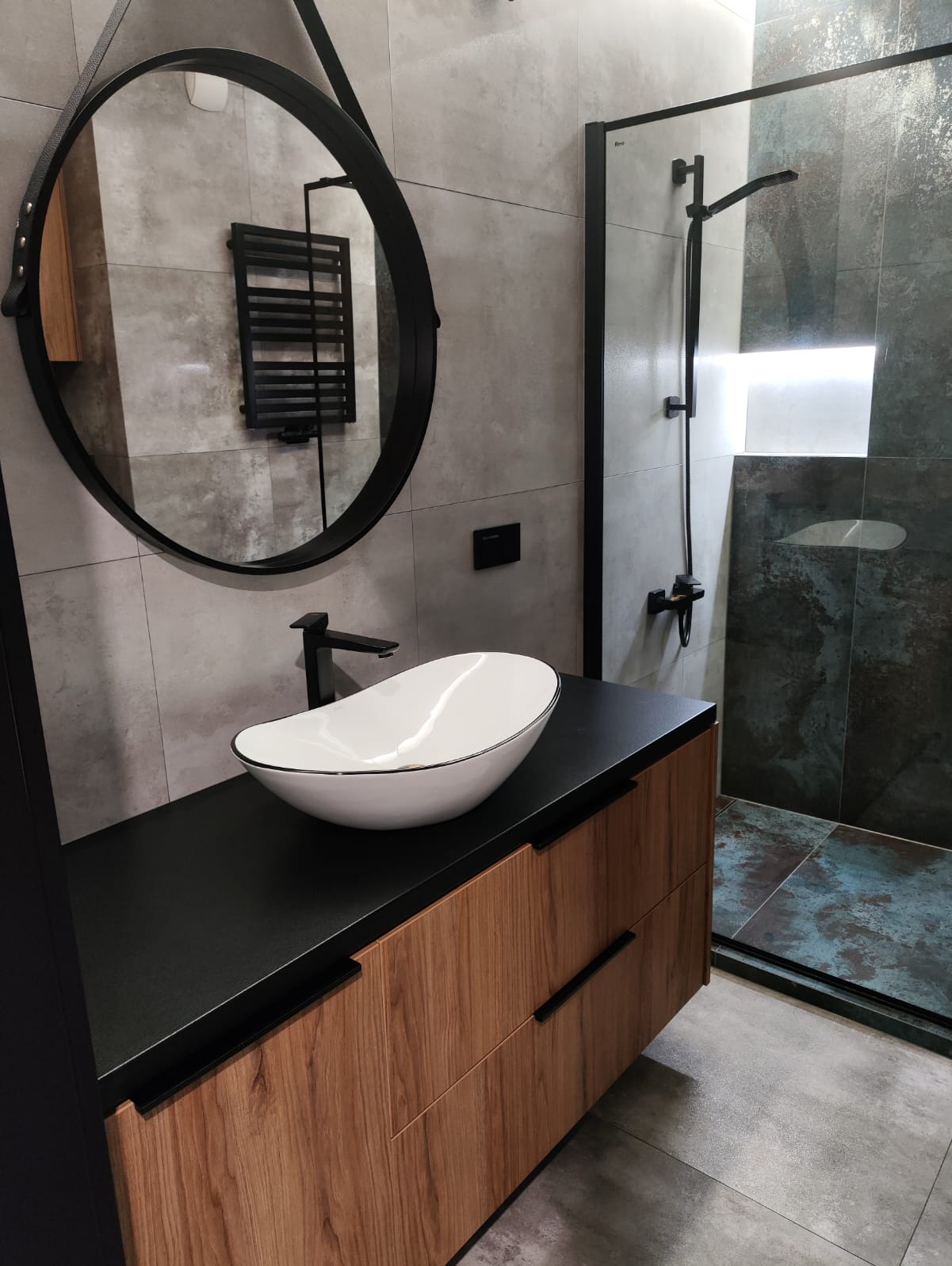 Nowoczesna łazienka: owalna umywalka na szafce z drewnianymi frontami, okrągłe lustro z czarnym obramowaniem i kabina prysznicowa z ciemnymi płytkami.
