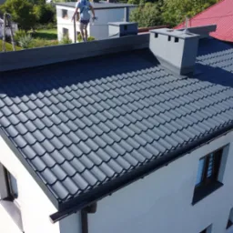 Nowy, ciemny dach z blachodach&oacute;wki na jasnym budynku, widok z g&oacute;ry. Kominy w kolorze dachu. Na dachu widoczna osoba w szelkach zabezpieczających.