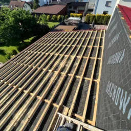 Konstrukcja drewniana dachu z membraną dachową w trakcie montażu, widok z góry na deskowanie i częściowo położone czerwone pokrycie dachowe.