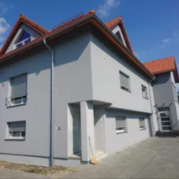 Elewacja budynku po malowaniu na szaro, z czerwonym dachem i szarymi roletami. Widoczny fragment chodnika z kostki brukowej i błękitne niebo.