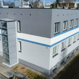 Nowoczesny budynek z przeszkloną windą zewnętrzną, pomalowany w odcieniach szarości z białym pasem i niebieskim akcentem. Ambulans zaparkowany w tle.