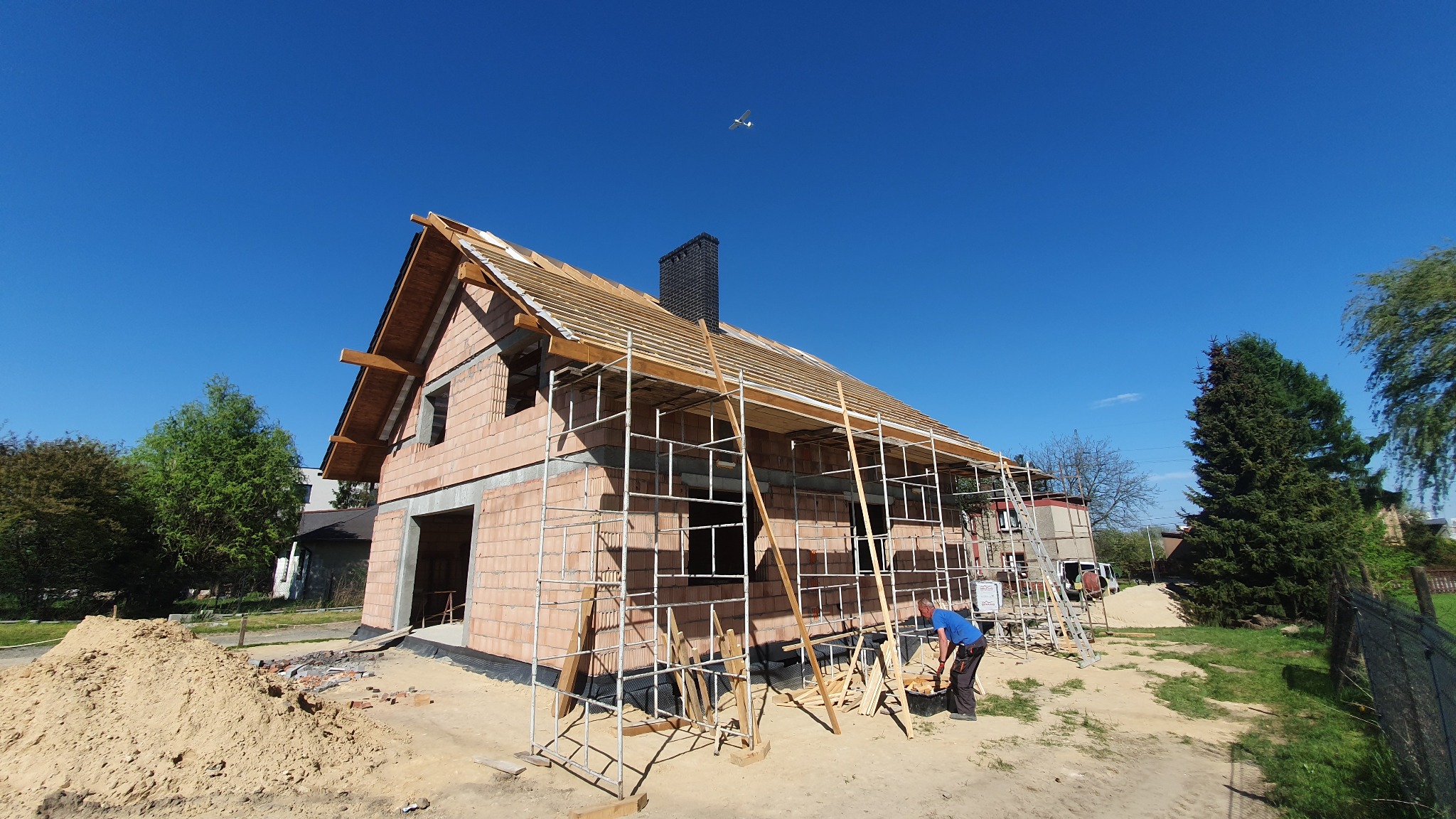Budowa domu jednorodzinnego z cegły, widoczne rusztowania, więźba dachowa w trakcie montażu, komin. Błękitne niebo z samolotem w tle, pracownik w dolnym rogu kadru.