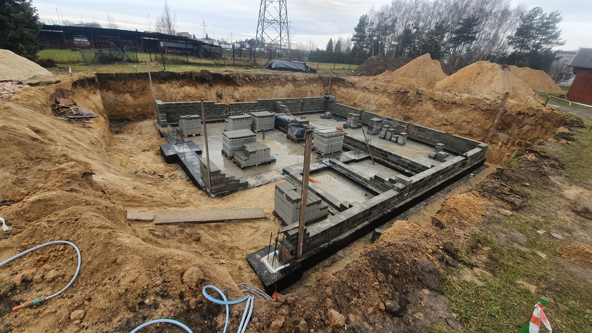 Fundamenty budynku z bloczków betonowych, częściowo zalane wodą, w wykopie otoczonym ziemią. Widoczne wystające pręty zbrojeniowe i ułożone bloczki. W tle drzewa i słupy energetyczne.