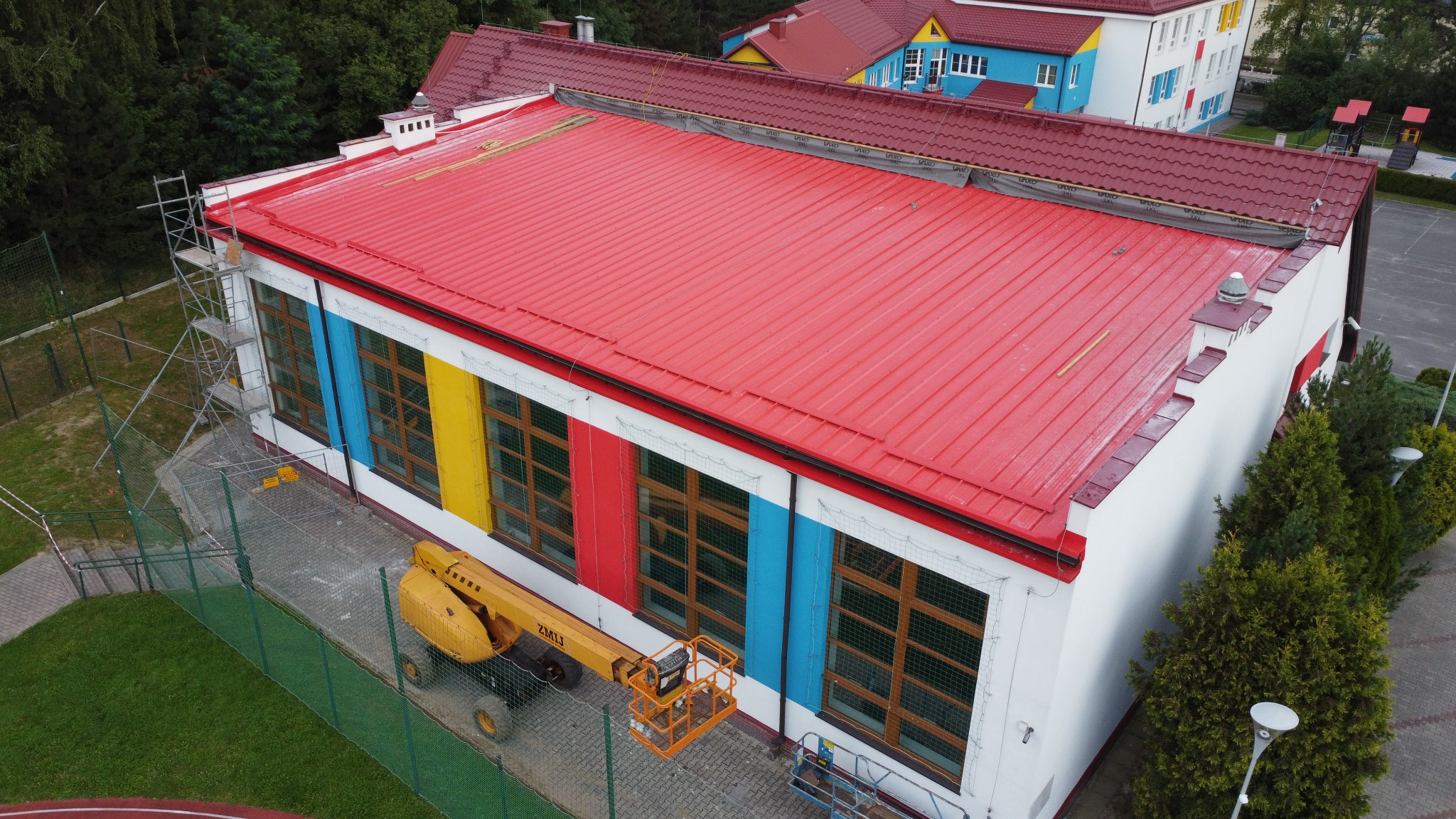 Czerwony dach budynku z kolorowymi akcentami (żółty, czerwony, niebieski) na ścianach. Podnośnik koszowy ZML13 przy ścianie. Widoczne rusztowania.