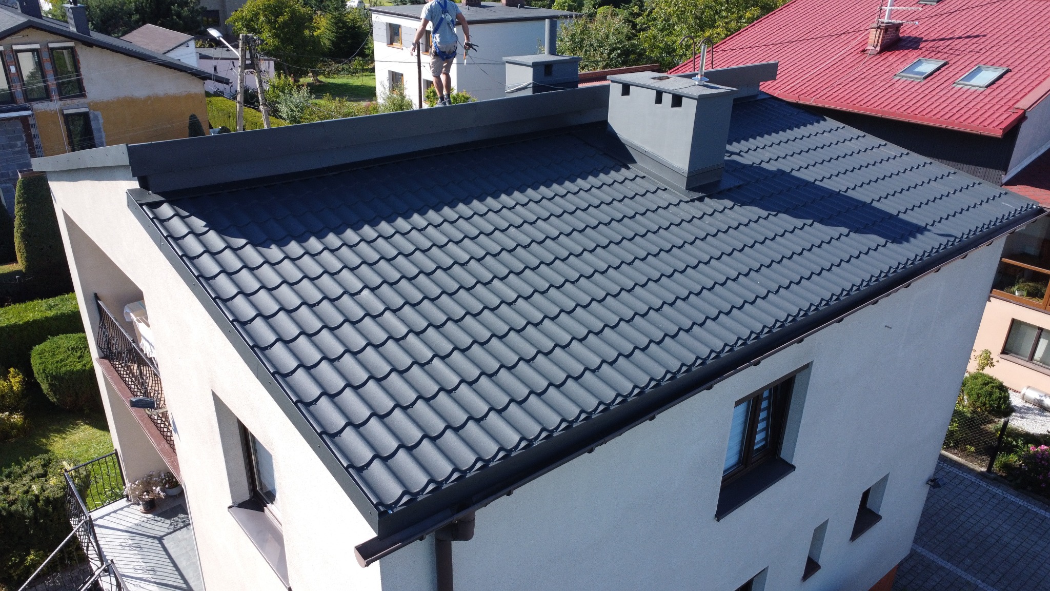Nowy, ciemny dach z blachodachówki na jasnym budynku, widok z góry. Kominy w kolorze dachu. Na dachu widoczna osoba w szelkach zabezpieczających.