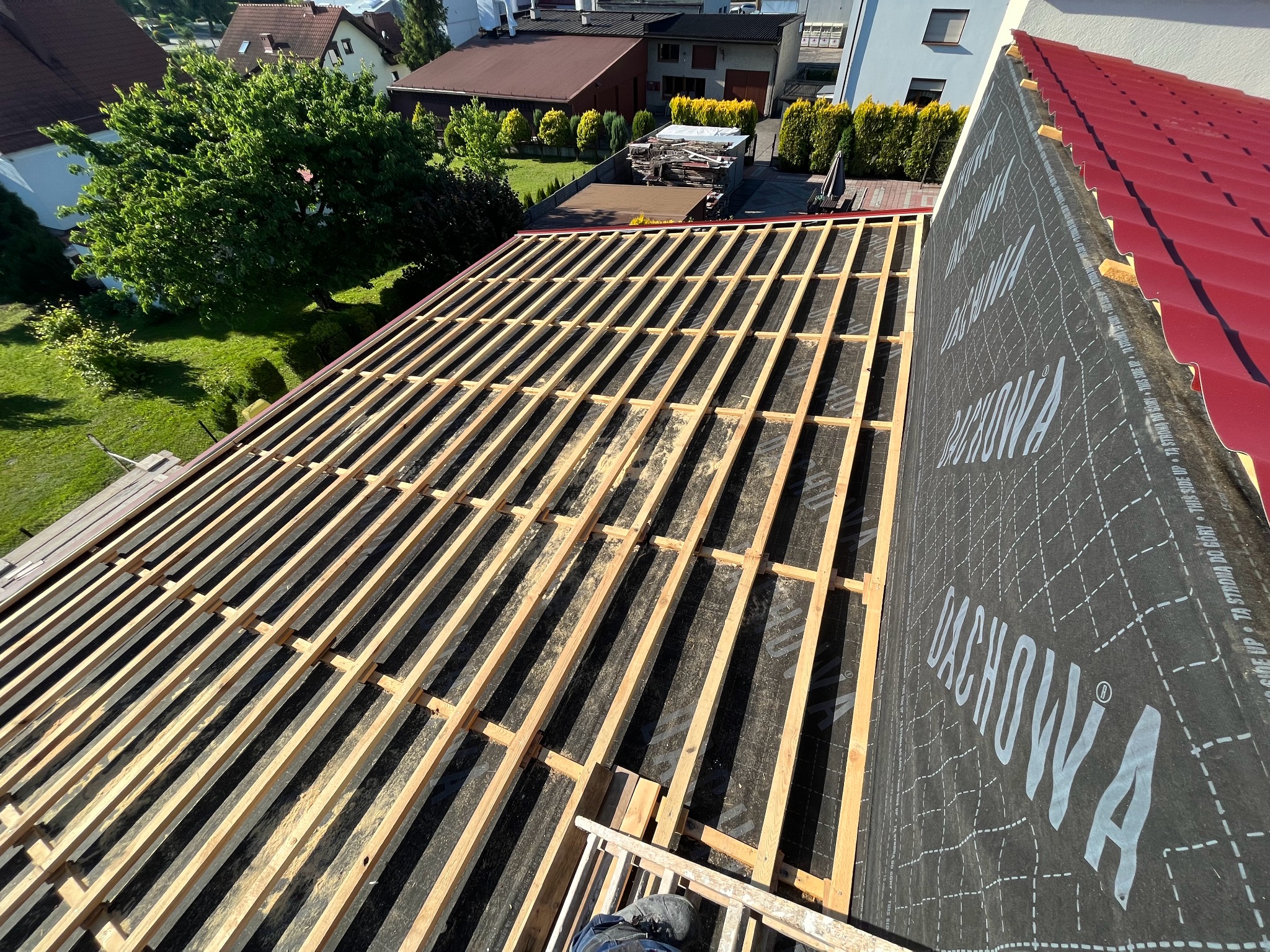 Konstrukcja drewniana dachu z membraną dachową w trakcie montażu, widok z góry na deskowanie i częściowo położone czerwone pokrycie dachowe.