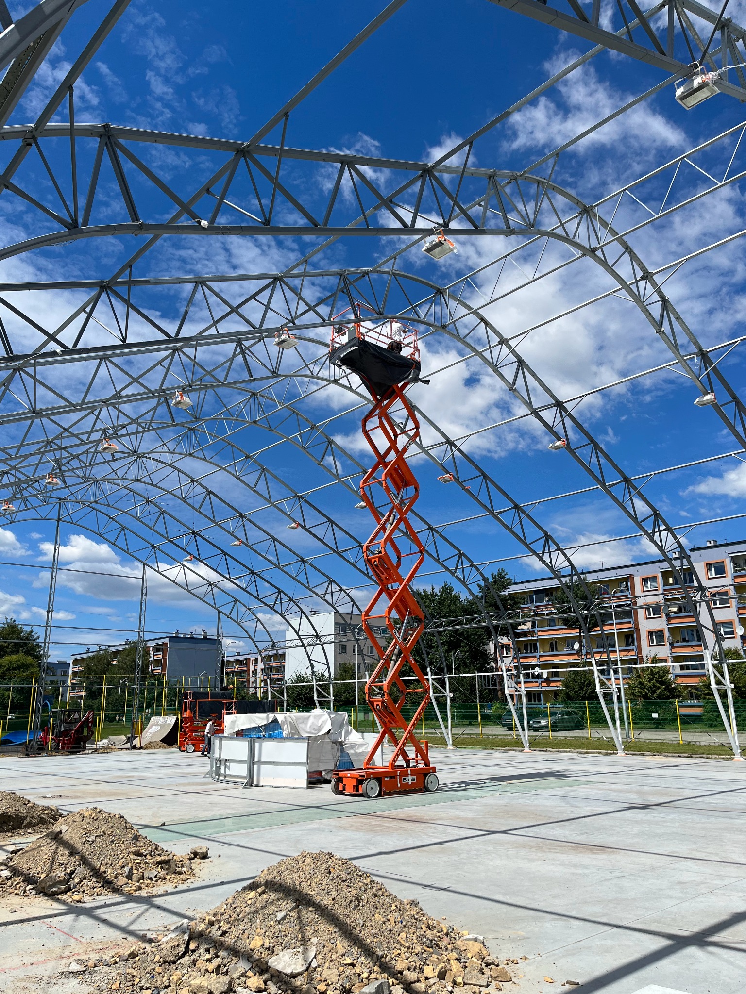 Konstrukcja stalowa hali w budowie, widok z dołu na niebo. Pomarańczowy podnośnik koszowy wewnątrz. Hałdy ziemi w tle. Budynki mieszkalne w oddali.