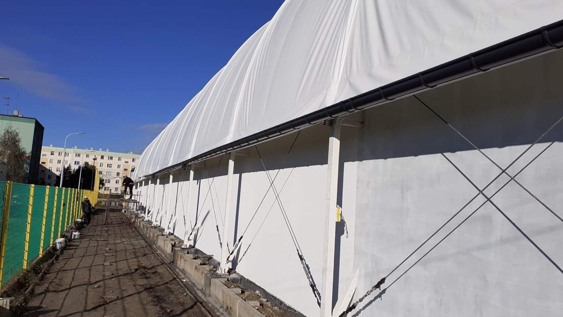 Biały, duży namiot halowy z czarną rynną, wsparty na słupach z odciągami. Widoczni pracownicy na rusztowaniu i zielone ogrodzenie. Błękitne niebo w tle.