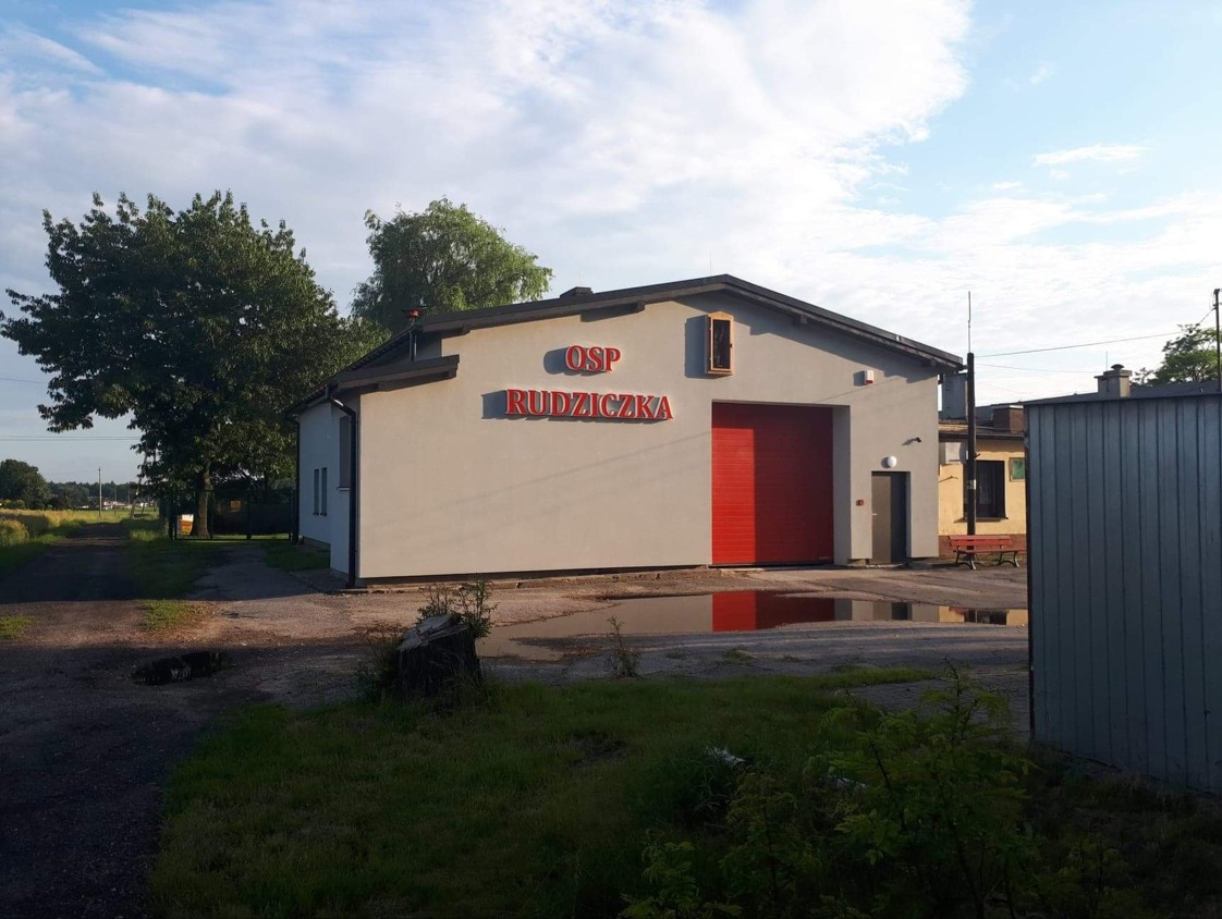 Budynek OSP Rudziczka z czerwoną bramą garażową i napisem na elewacji, otoczony zielenią i kałużami. Widoczne drzewa i fragment drogi.