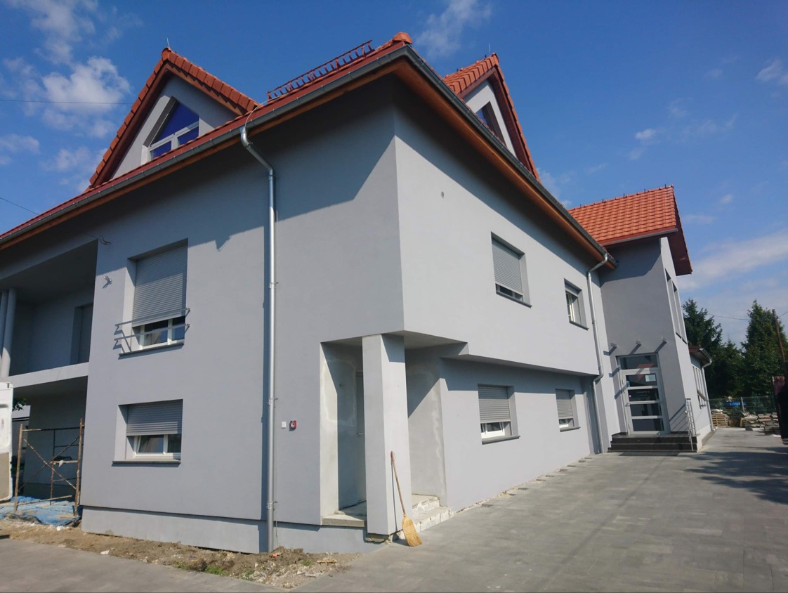 Elewacja budynku po malowaniu na szaro, z czerwonym dachem i szarymi roletami. Widoczny fragment chodnika z kostki brukowej i błękitne niebo.