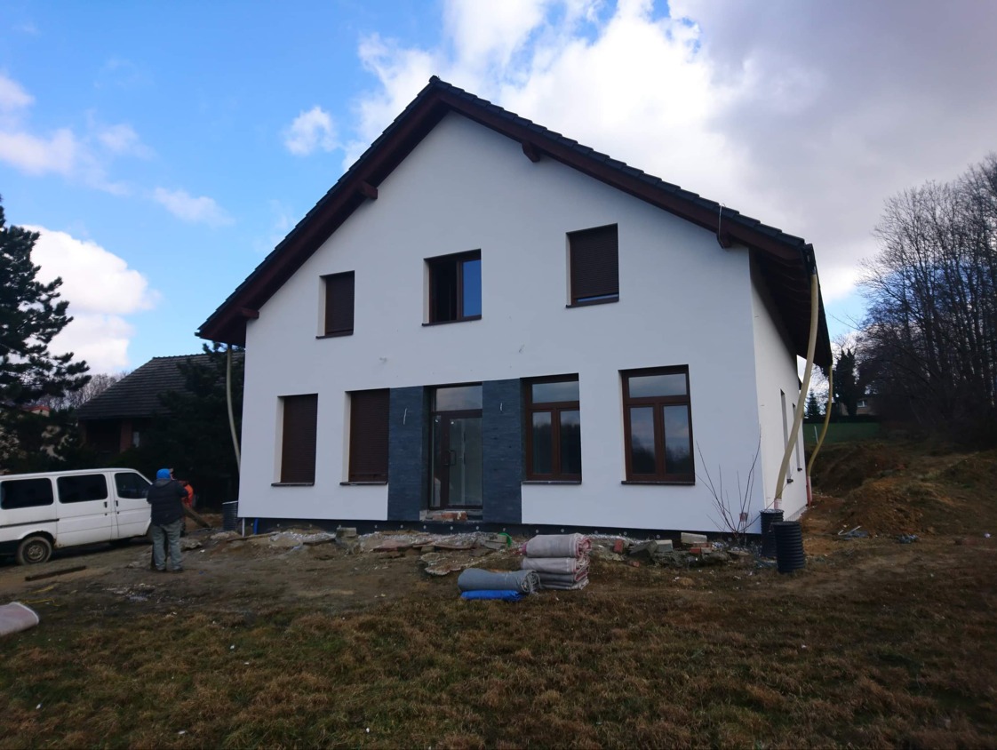 Biały dom z brązowymi oknami i roletami w trakcie prac wykończeniowych. Widoczne materiały budowlane i pracownicy. Tło: pochmurne niebo i drzewa.