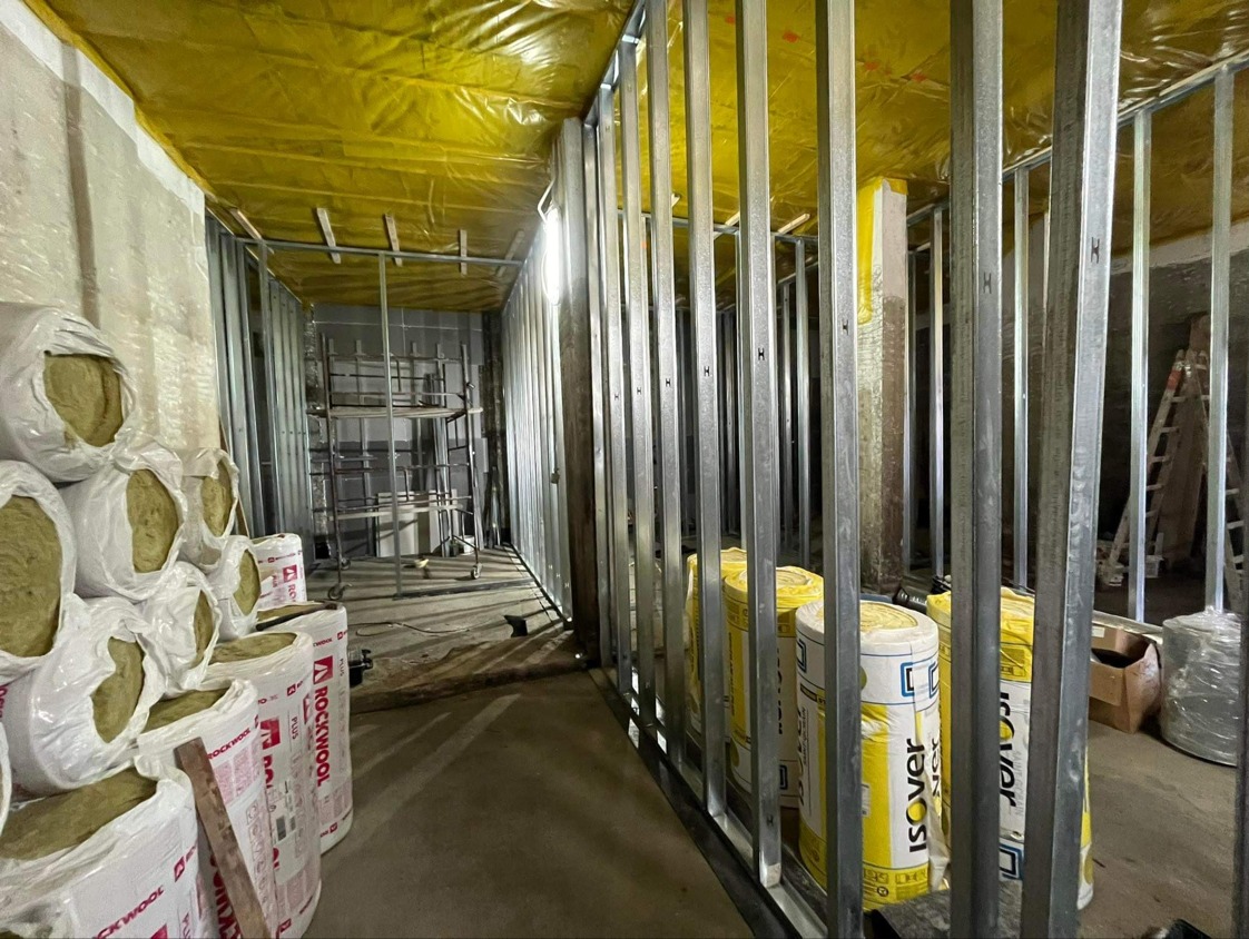 Wnętrze w trakcie prac wykończeniowych: stalowa konstrukcja szkieletowa, izolacja Rockwool i Isover, żółta folia na suficie, rusztowanie w głębi pomieszczenia.