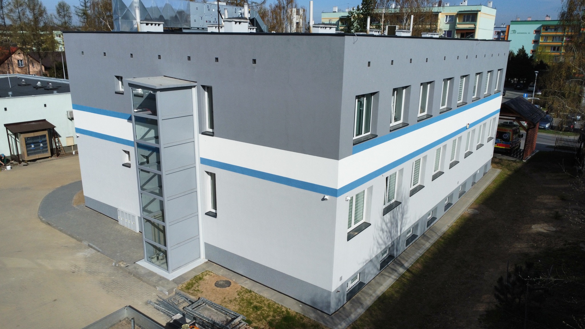 Nowoczesny budynek z przeszkloną windą zewnętrzną, pomalowany w odcieniach szarości z białym pasem i niebieskim akcentem. Ambulans zaparkowany w tle.