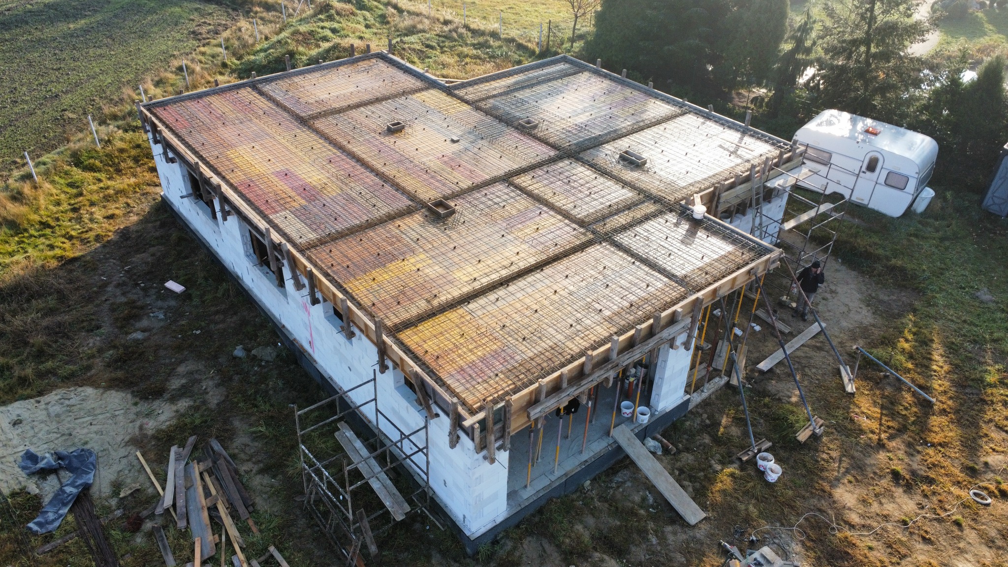 Budowa domu jednorodzinnego: widok z góry na strop żelbetowy z szalunkiem i zbrojeniem, bloczki fundamentowe, rusztowania. Przyczepa kempingowa w tle.