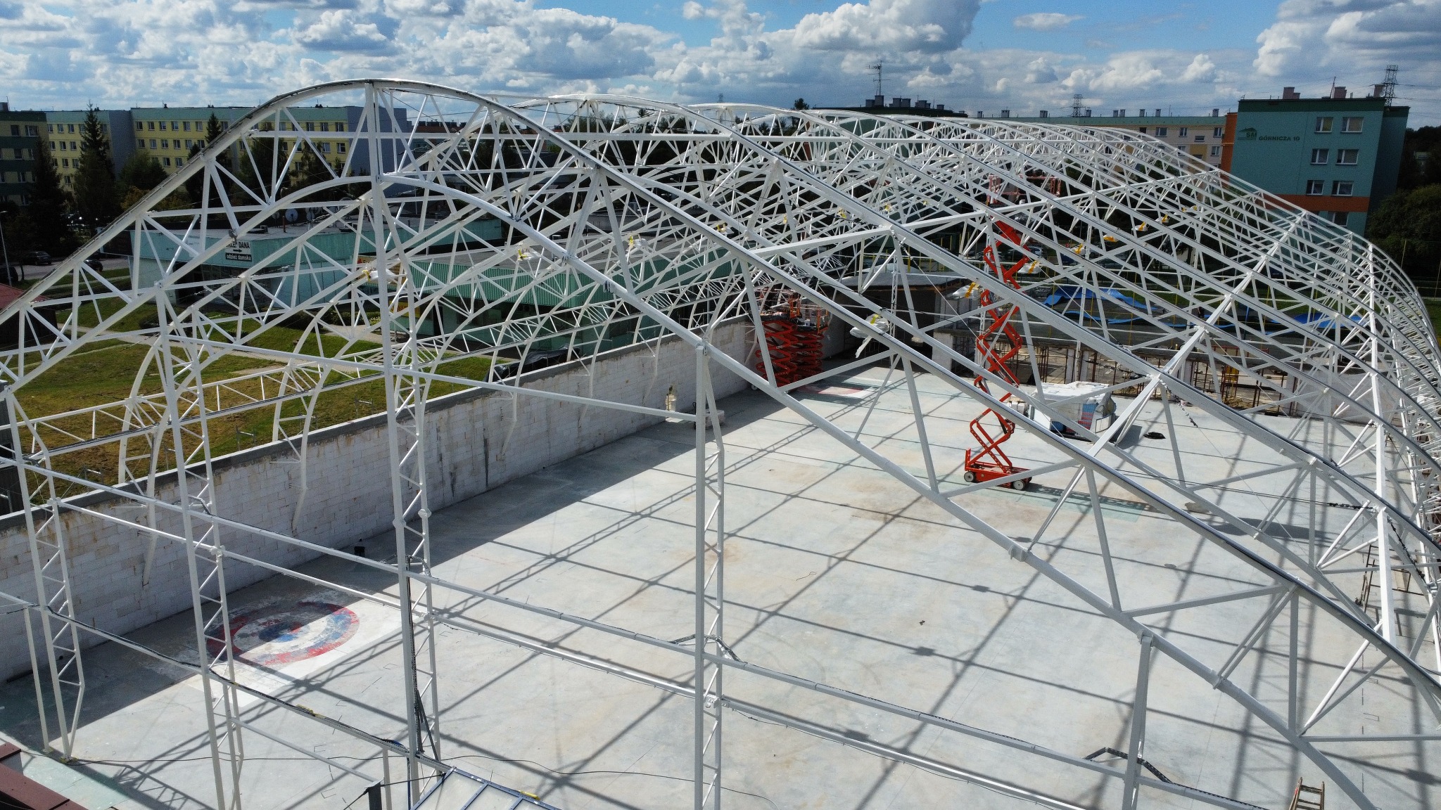 Konstrukcja stalowa hali sportowej w trakcie budowy, widok z góry. Białe elementy tworzą łukowatą kratownicę na tle błękitnego nieba i bloków mieszkalnych.