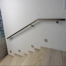 Nowoczesna balustrada schodowa z metalu, mocowana do ściany z dekoracyjną tapetą. Schody wykończone drewnem w jasnym odcieniu. Minimalistyczny design, oświetlenie punktowe.