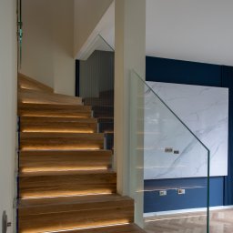 MAKRO GLASS ROBERT KUDŁA - Drewniane schody z podświetleniem LED i szklaną balustradą, widok z dołu. W tle ściana w kolorze granatowym z dekoracyjnym panelem imitującym marmur.