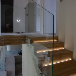 MAKRO GLASS ROBERT KUDŁA - Szklana balustrada schodowa z podświetleniem LED, widok z góry. Schody drewniane, jasne wnętrze z widocznymi drzwiami i fragmentem kuchni w tle.