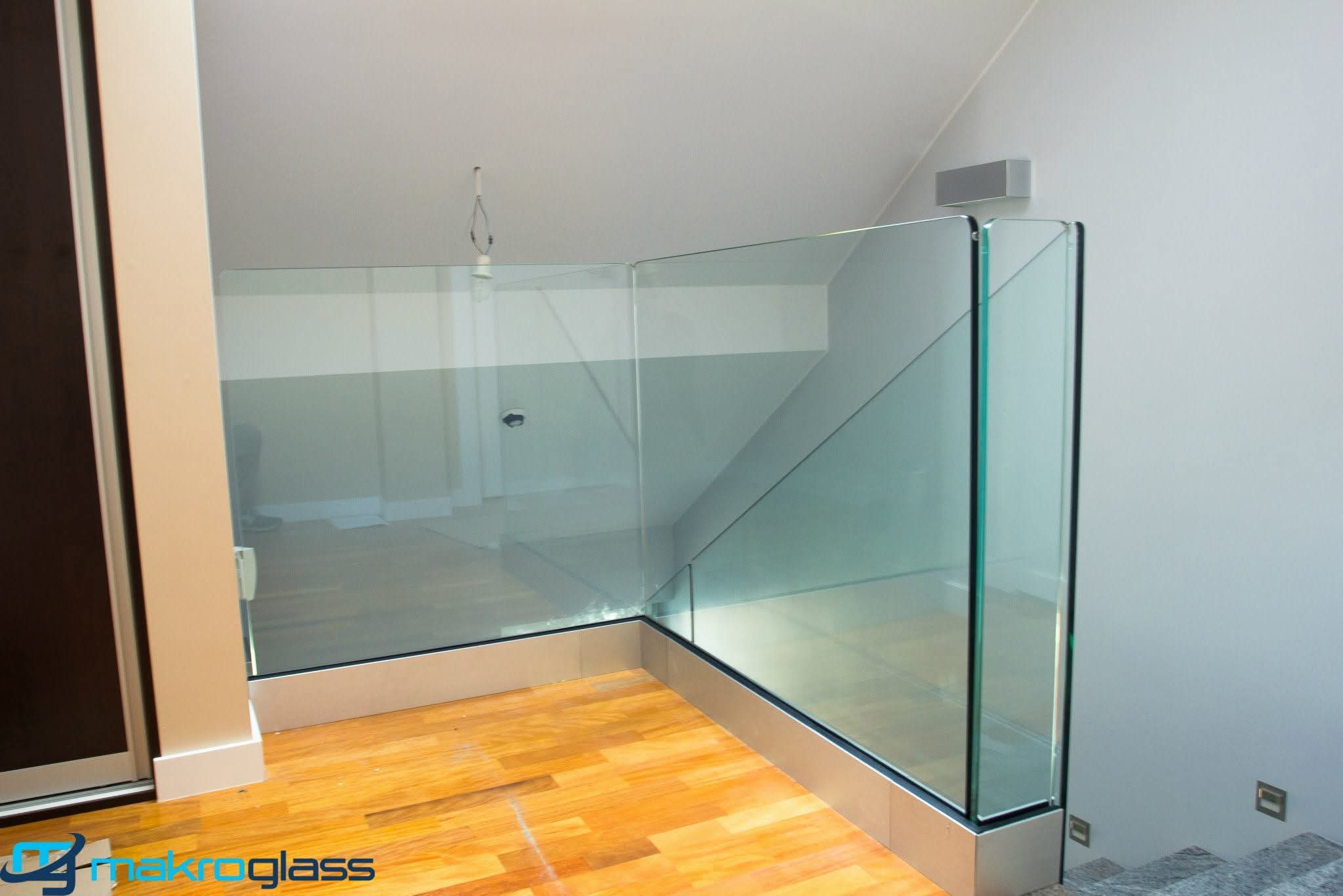 Szklana balustrada schodowa z metalową ramą, widoczna z perspektywy podłogi z drewnianych paneli. Minimalistyczny design, jasne wnętrze z szarą ścianą.