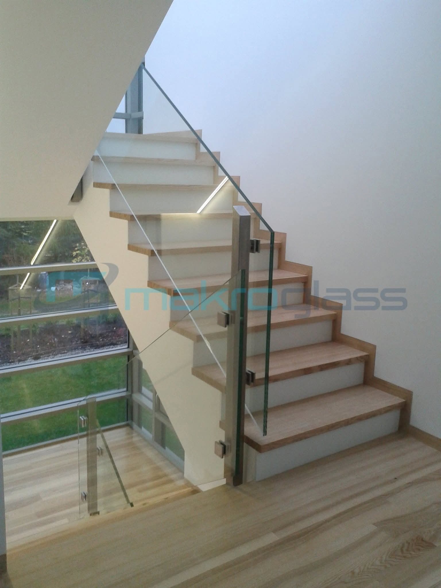 Nowoczesne schody z drewnianymi stopniami i szklaną balustradą, wspartą na metalowych słupkach. Widok z dołu, z oknem w tle.