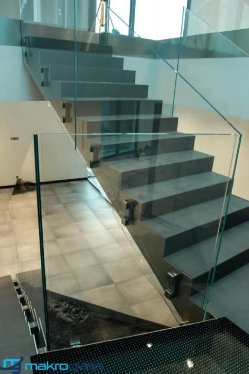 Nowoczesne schody z szarymi stopniami i szklaną balustradą, zabezpieczoną metalowymi uchwytami. Minimalistyczny design z widocznym logo firmy w dolnym rogu.
