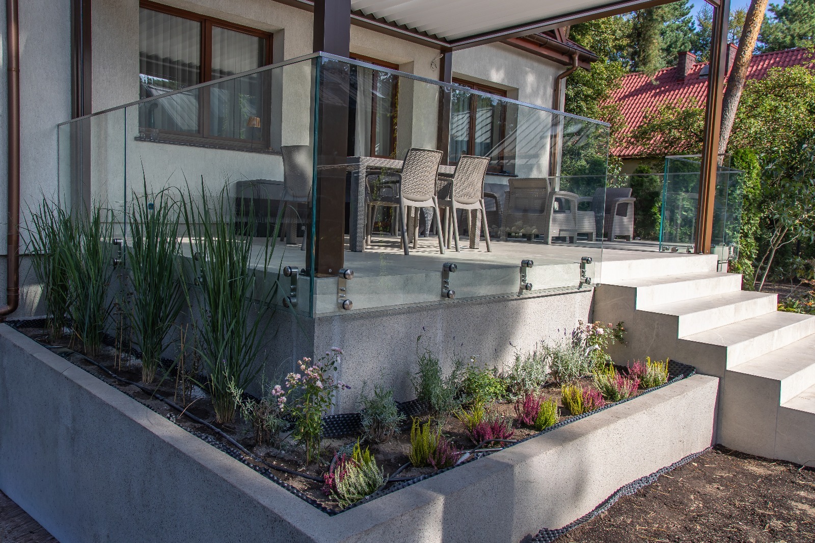 Szklana balustrada na tarasie z roślinami w donicy. Widoczne szklane panele i mocowania. Na tarasie meble ogrodowe. Tło: dom z ogrodem.