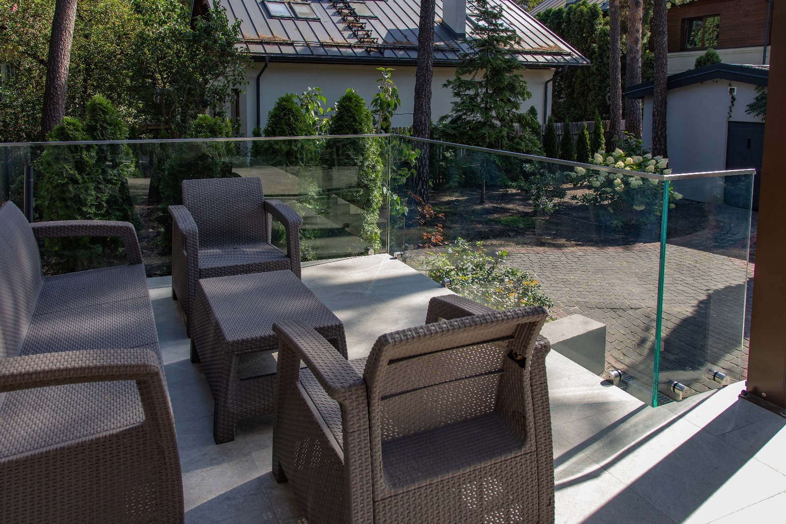 Szklana balustrada tarasowa z brązowymi meblami ogrodowymi na tle zieleni i domu z szarą dachówką. Widoczne detale mocowania szkła.