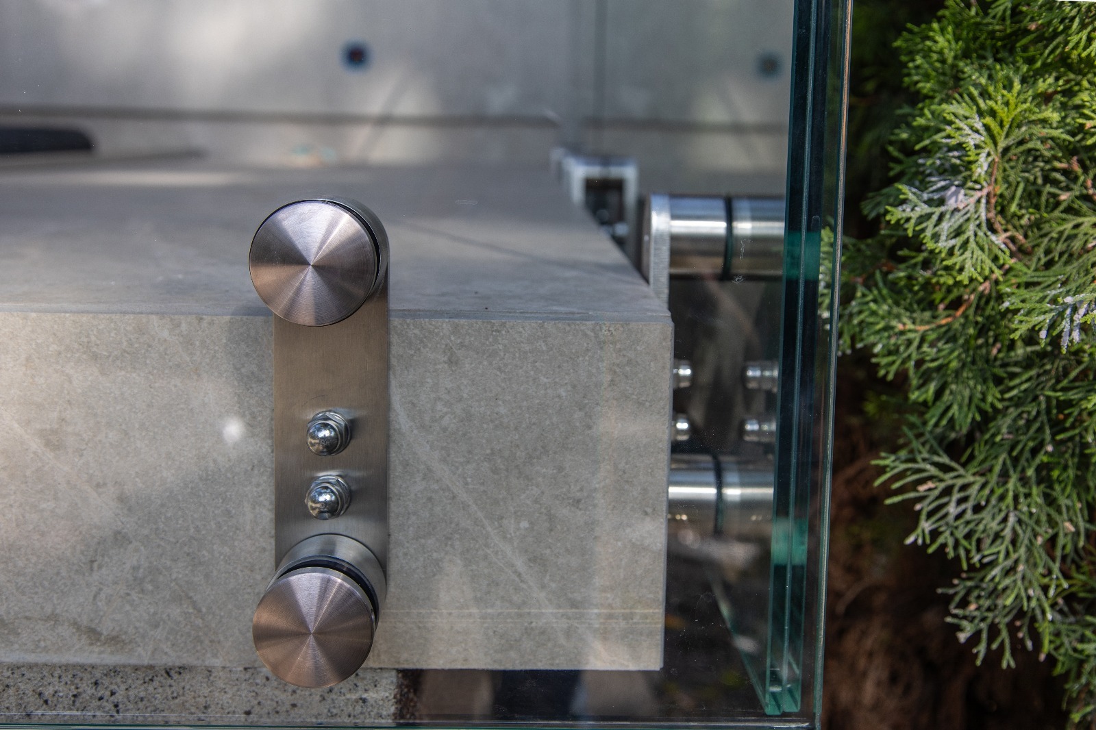 Szklana balustrada z detalami mocowań, widoczne metalowe śruby i okrągłe uchwyty, obok zieleń. Precyzja wykonania i nowoczesny design.