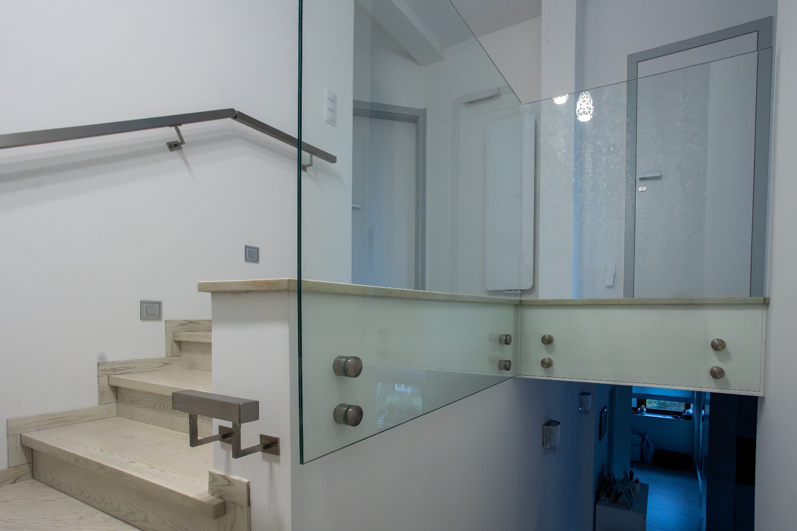 Nowoczesna szklana balustrada na schodach z metalową poręczą, mocowana punktowo. Widok z dołu na górę, wnętrze domu z białymi ścianami i drzwiami.