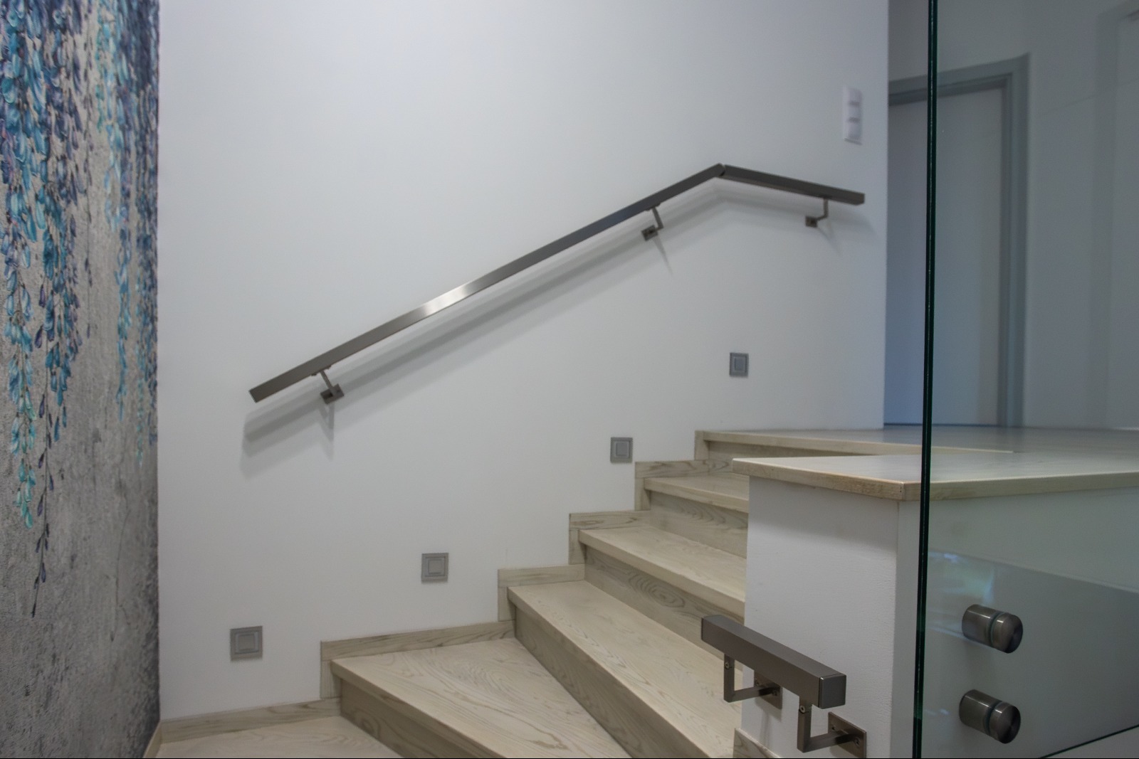 Nowoczesne schody z drewnianymi stopniami i metalową balustradą, ozdobione szklanym panelem. Ściana z wzorem kwiatowym po lewej stronie.