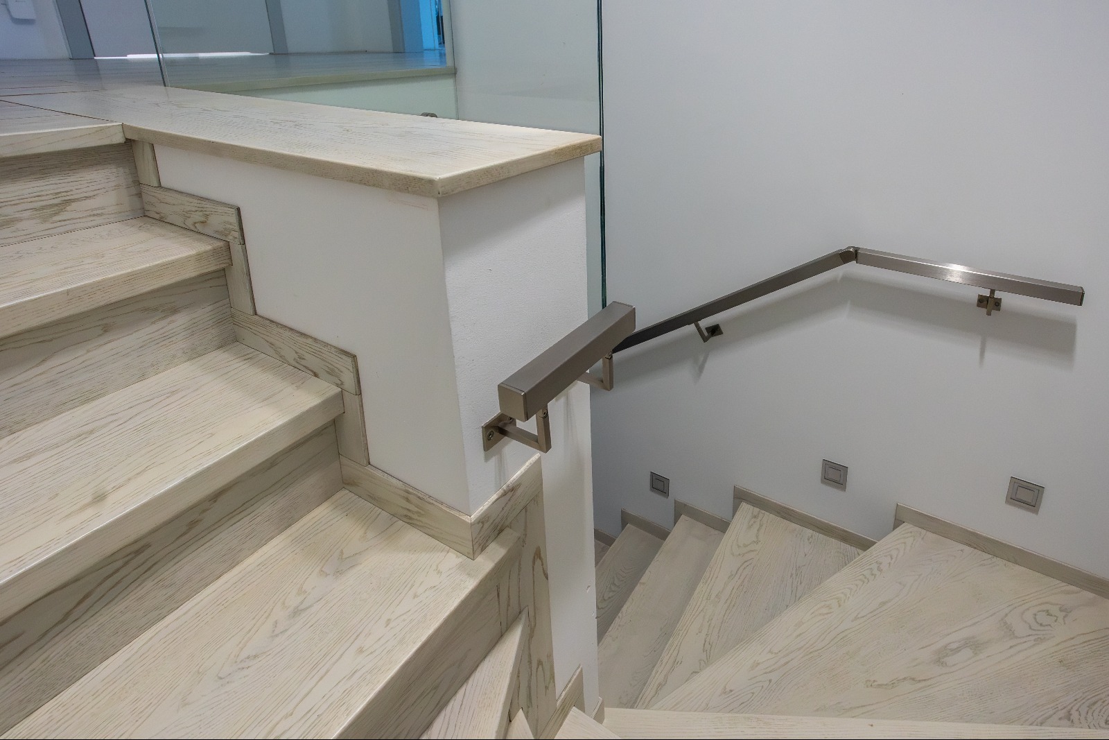 Jasne, drewniane schody z metalową poręczą zamocowaną do ściany i szklaną balustradą. Minimalistyczny design, nowoczesne wykończenie.