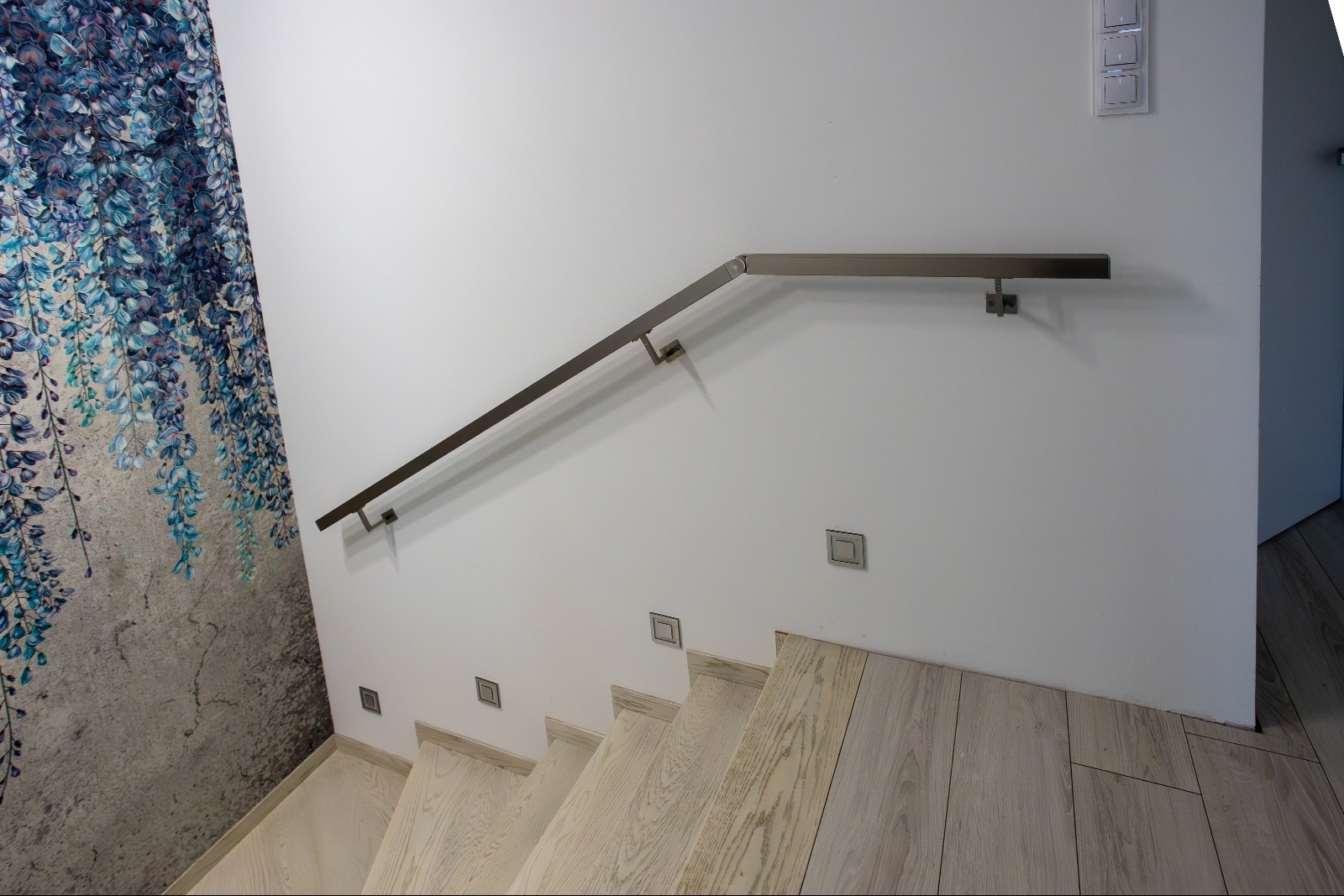 Nowoczesna balustrada schodowa z metalu, mocowana do ściany z dekoracyjną tapetą. Schody wykończone drewnem w jasnym odcieniu. Minimalistyczny design, oświetlenie punktowe.