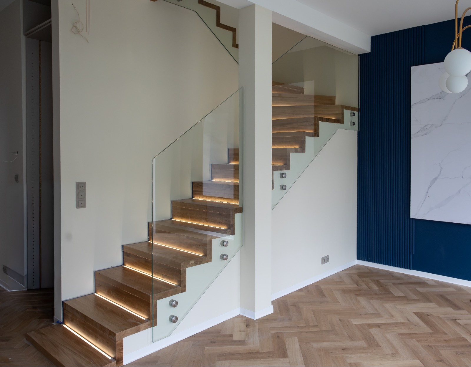 Nowoczesne schody z drewnianymi stopniami podświetlonymi LED, zabezpieczone szklaną balustradą. Jasne ściany i parkiet w jodełkę. Elementy dekoracyjne na ścianie.