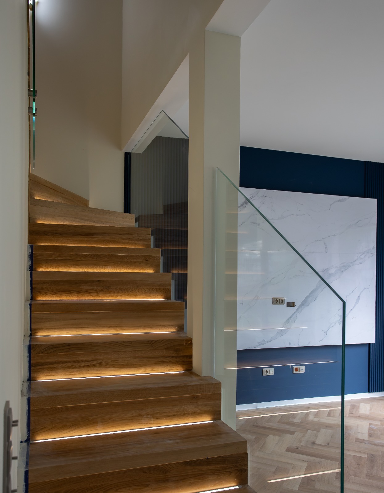 Drewniane schody z podświetleniem LED i szklaną balustradą, widok z dołu. W tle ściana w kolorze granatowym z dekoracyjnym panelem imitującym marmur.