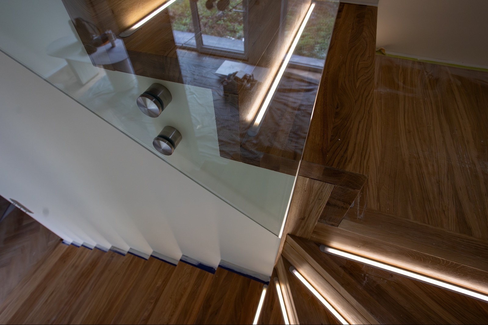 Szklana balustrada schodowa z oświetleniem LED, mocowana na metalowych uchwytach. Widok z góry na drewniane schody z jasnego drewna.