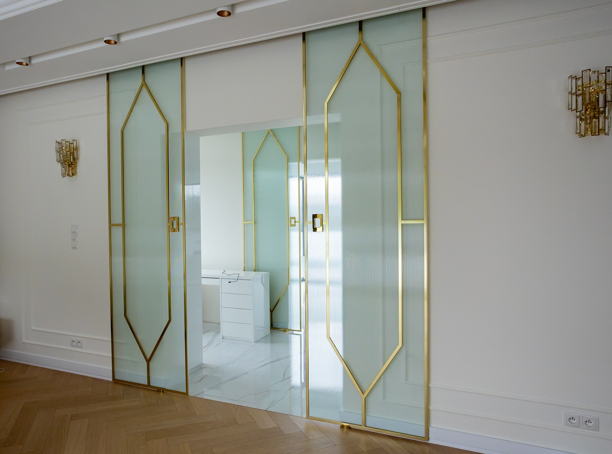 Eleganckie, przesuwne drzwi szklane z geometrycznym, złotym obramowaniem w jasnym wnętrzu. Widoczny parkiet i marmurowe płytki podłogowe.